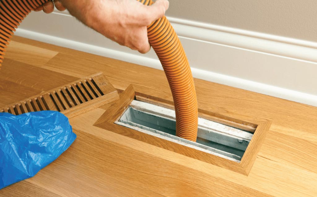 Technician using a hose to clean a floor air duct, highlighting the benefits of air duct cleaning for improved indoor air quality, energy efficiency, and a cleaner living environment.