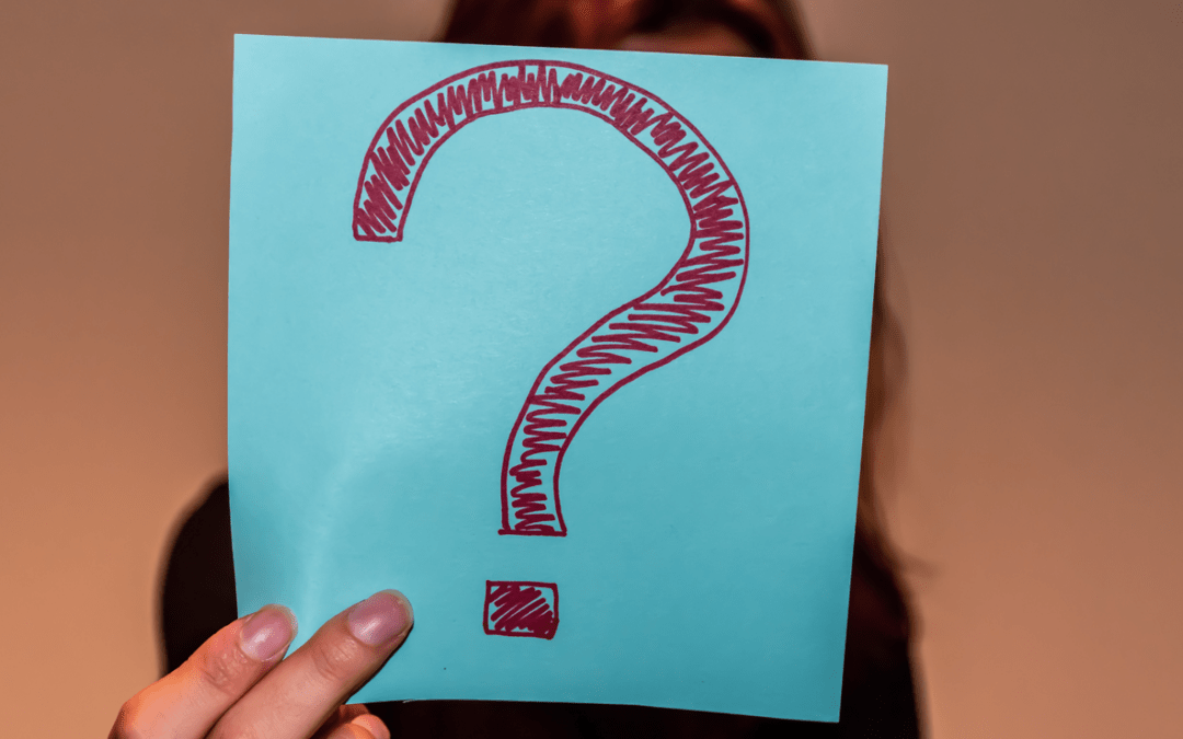 A close-up shot of an unrecognizable young Caucasian redhead woman's hand holding a light blue paper sheet with a hand-drawn question mark and hiding her face behind it